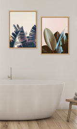 Two framed botanical prints above a white freestanding bathtub, depicting dark green and pink-tinted ficus leaves on soft pink backgrounds.