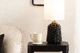 Black woven rattan table lamp with a white cylindrical shade on a dark wood side table next to a white ceramic mug and light-colored textured wall.
