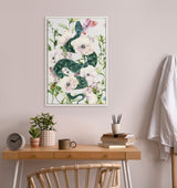 Framed canvas print of a green snake weaving through large pale pink flowers and green foliage against a white background, hanging above a wooden desk with plants and books.