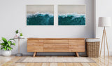 Two framed canvases showing aerial views of ocean waves crashing with blue, white, and sandy tones, displayed above a wooden mid-century modern sideboard in a minimalist room.