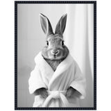 Black and white portrait of a rabbit standing upright wearing a white bathrobe tied at the waist, with a neutral curtain backdrop and a dark textured frame.