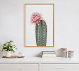 Vertical canvas print of a green cactus with a large pink flower on top, framed in light wood and hanging above a white cabinet with minimal decor.