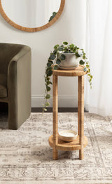 Tall, round plant stand made of light-colored woven material, holding a cascading green potted plant and a small white bowl below, set on a patterned rug near an olive-green sofa.