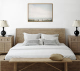 Minimalist bedroom with a light wood bedframe, white bedding, two matching wooden nightstands with black lamps, and a pale sky beach scene framed canvas centered above the bed.