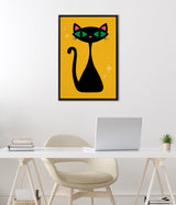 Black stylized cat with green eyes and pink inner ears on a mustard yellow background with small star shapes, framed and hung above a modern white desk with laptop and plant.