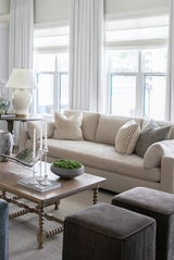 Light beige sofa with patterned greige pillows, positioned by tall windows with white curtains, next to a wooden coffee table with white candleholders and a green centerpiece.