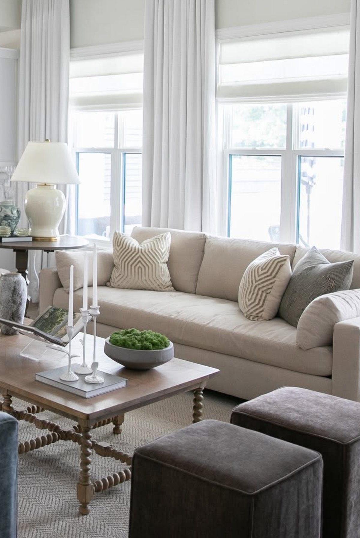 Light beige sofa with patterned greige pillows, positioned by tall windows with white curtains, next to a wooden coffee table with white candleholders and a green centerpiece.