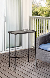 Slim black metal side table with perforated top and crossbar support, standing on a wooden deck beside a woven chair with a blue cushion, holding a glass of water.