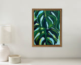 Framed canvas painting of large green monstera leaves with white and blue accents against a light background, hanging on a light gray wall above a beige surface.