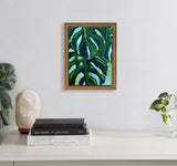 Framed 8x10 painting of large green monstera leaves with dark and light green hues on a light blue background, hanging above a white desk with plants and stacked books.