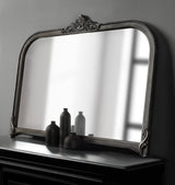 Arched wall mirror with a dark, ornate frame featuring beaded detailing and a decorative crest, reflecting a blurred window and four matte black decorative bottles below.