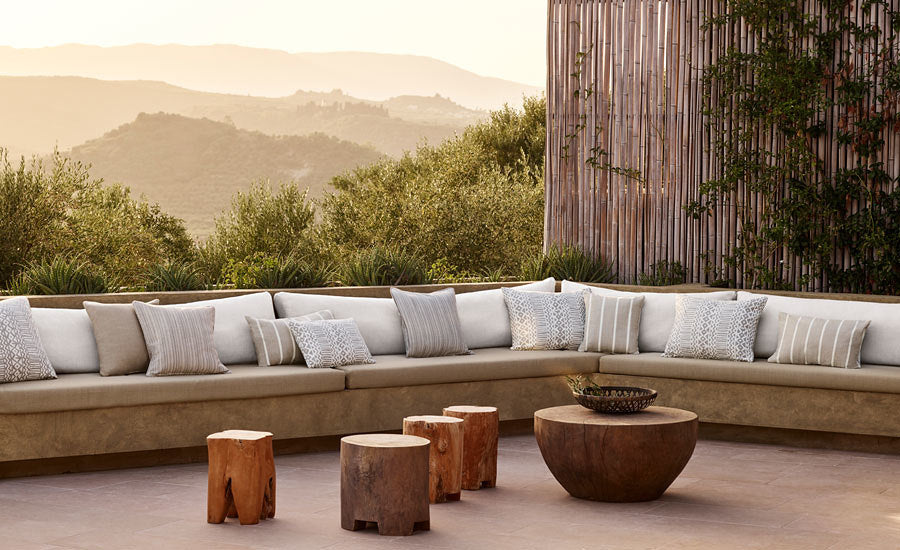 Outdoor curved beige sectional sofa with multiple grey and patterned cushions, arranged around round wooden stools and a circular wooden coffee table, with a bamboo screen and mountain view in the background.