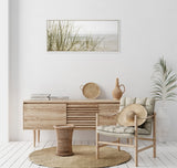 Neutral-toned room with natural wood sideboard, cushioned wooden chair holding a straw hat, rattan stool on woven rug, and framed beach grass canvas overhead.