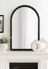 Arched wall mirror with black frame featuring rounded bead details along the edge, displayed on a white mantel with sculptural white ceramic vases and a textured pot holding greenery.