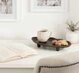 Oval dark wood tray with short legs holding a white mug and three pastries, positioned on a white desk beside an open book and a vase with small branches