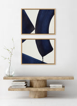 Two framed abstract canvases with bold navy blue geometric shapes on white backgrounds, arranged vertically above a minimalist wooden console table with books and a glass vase.