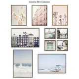 Nine framed artworks featuring soft pink flowers, Parisian buildings, palm trees, Amsterdam houses, Eiffel Tower view, a green door, and ocean scenes with waves and lifeguard tower.