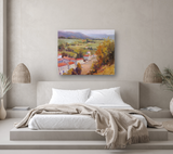 Impressionistic canvas print of a French village with red-roofed houses, green fields, and distant mountains, hung above a modern bed with beige bedding.