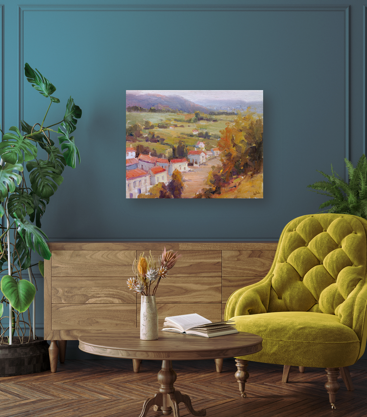 Impressionist-style canvas painting of a French village with red rooftops, green fields, and distant hills, hanging on a teal wall above a wooden sideboard.