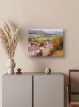 Impressionist-style canvas painting of a French village with red-roofed houses, green fields, and distant mountains, displayed on a beige wall above a minimalist cabinet.