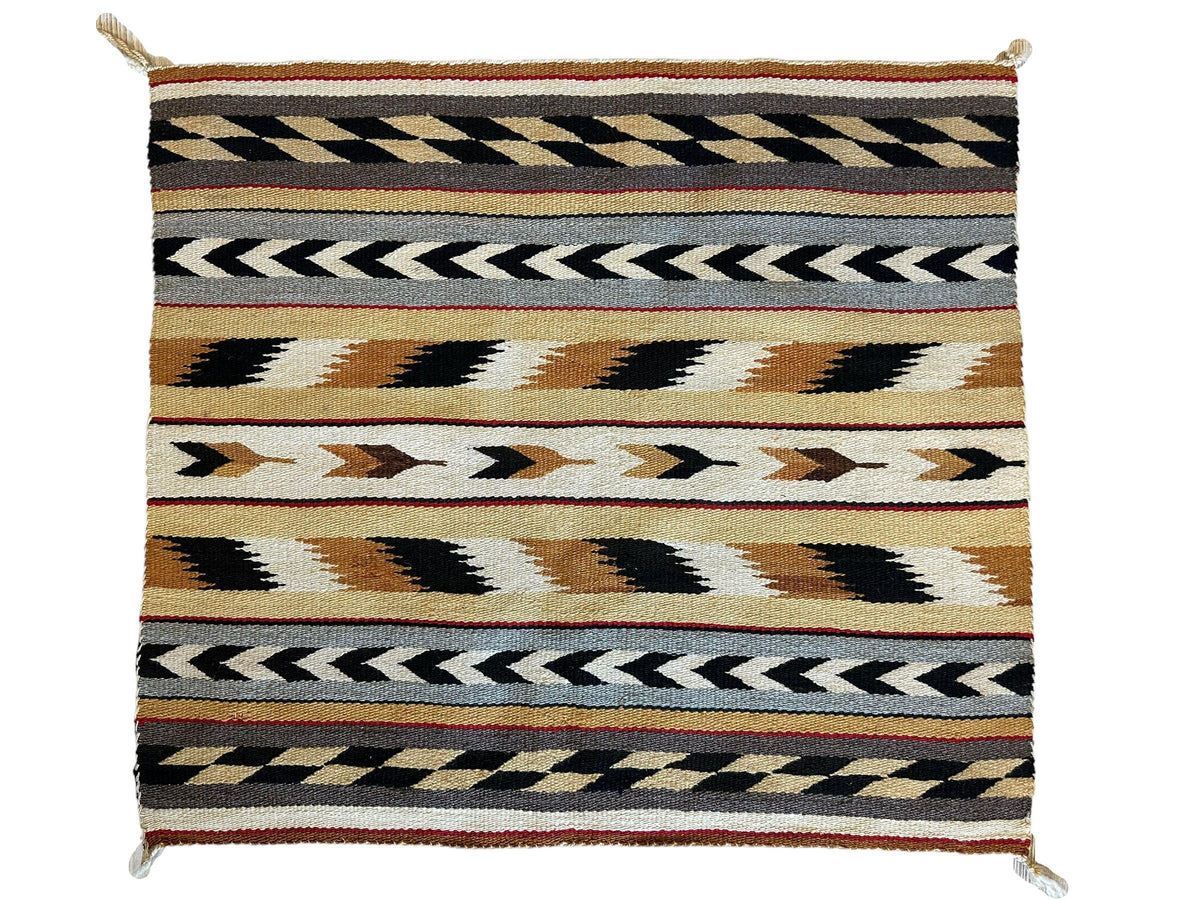 Rectangular vintage Navajo rug with horizontal geometric patterns in black, white, tan, and rust colors featuring chevrons and zigzags, with knotted tassels at corners.