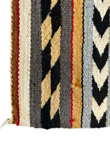 Close-up of vintage Navajo rug corner with vertical stripes in beige, black, gray, and red, featuring zigzag and diagonal woven patterns on textured wool fabric.