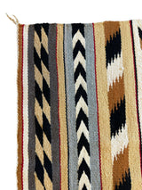 Section of a Native American Navajo rug with vertical zigzag stripes in black, beige, white, gray, and brown on a tan background.