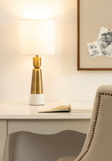 Table lamp with white cylindrical fabric shade and base of white marble topped by brushed brass, sitting on white wooden desk with open book and beige studded chair.