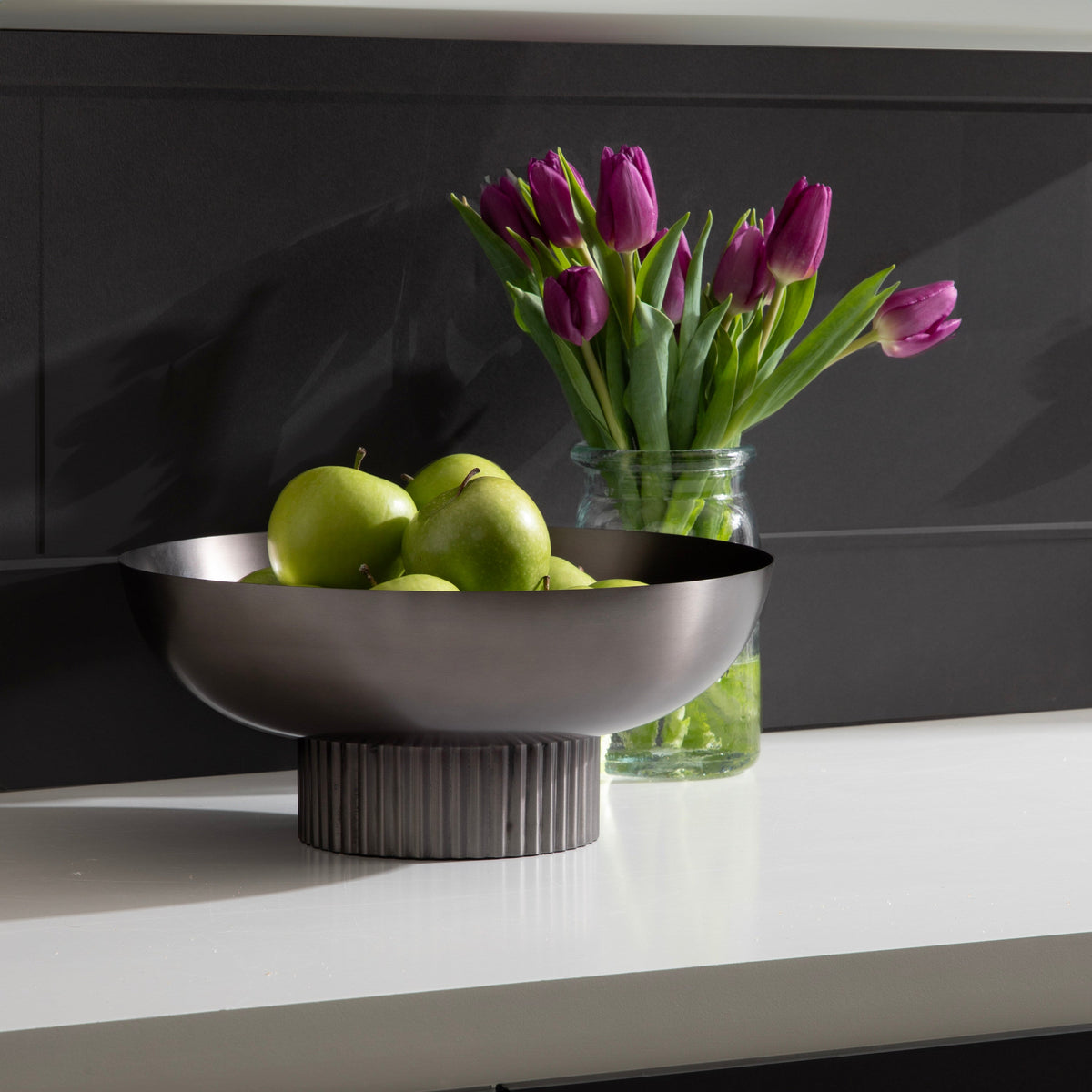 Matte dark gray metal bowl with vertical ribbing on its cylindrical base filled with green apples, next to a glass jar of purple tulips on a white surface against a dark background.