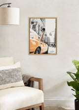 Framed canvas print of a giraffe with sunglasses poking out of an orange New York taxi in a black-and-white city street scene, hung on a light wall above a beige armchair and near a green plant.