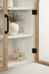 White wall cabinet with glass doors featuring light wood frames and black metal handles, showing a white vase with branches and two decorative glass bottles inside.