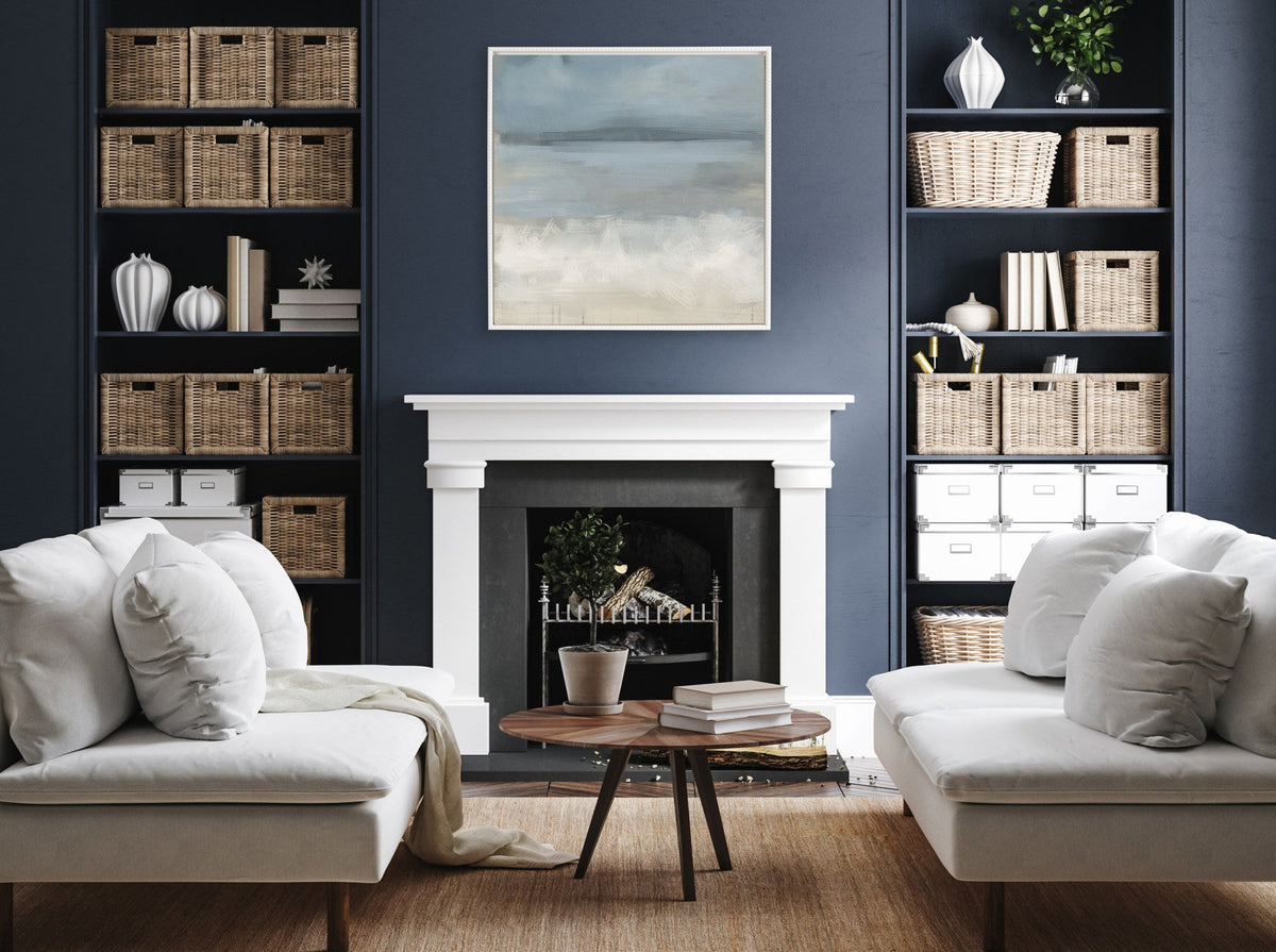Contemporary living room with two beige sofas facing each other, a round wooden coffee table, blue-gray walls, built-in shelves with wicker baskets, and an abstract landscape painting above a white fireplace.