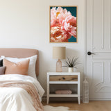 Framed canvas of soft pink peonies against a dark teal background hanging above a white bedside table with a beige lamp and a small potted plant in a light-toned bedroom.