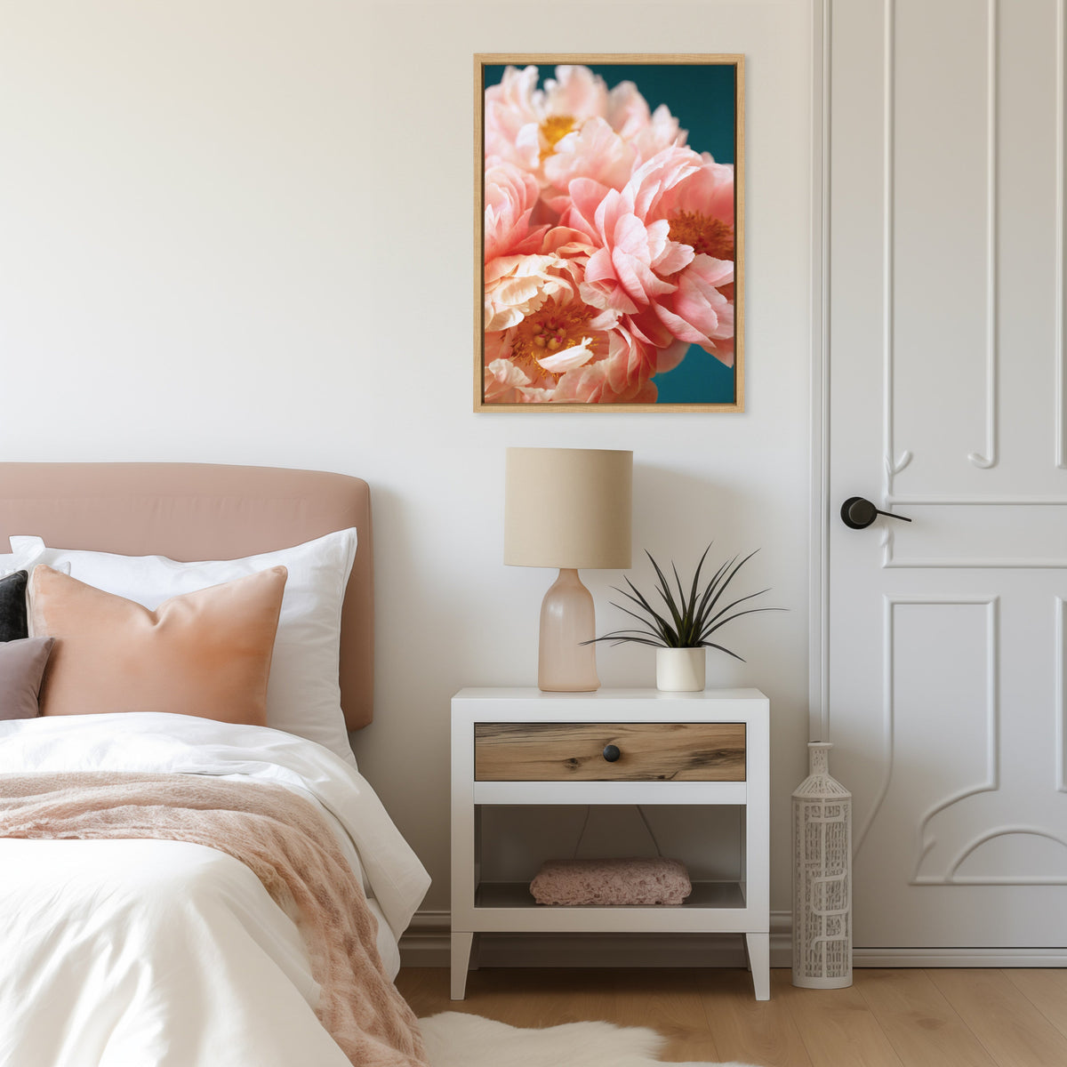 Framed canvas of soft pink peonies against a dark teal background hanging above a white bedside table with a beige lamp and a small potted plant in a light-toned bedroom.