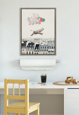 Framed canvas print above a desk depicting a gray elephant lifted by pastel-colored balloons over Paris rooftops in a minimalist room with light wooden furniture and art supplies.