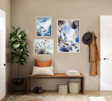 Three framed abstract art pieces in blue, gray, and beige tones arranged on a beige wall above a wooden bench with a decorative pillow, books, and baskets below.