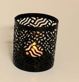 Black metal votive candle holder with a wavy cutout pattern, showing a lit candle inside casting warm light through the shapes.