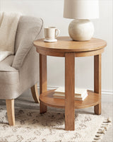 Round wooden side table with a smooth top and lower shelf holding stacked books, positioned beside a gray upholstered armchair with a textured throw.
