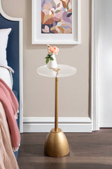 Round side table with a clear glass top and a matte gold pedestal base, holding a small white vase with pink flowers, set against a beige wall with framed floral art.