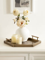 Hexagon-shaped bronze metal tray holding a white ceramic vase with cream-colored roses and two ivory flameless candles on a white cabinet