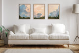 Triptych of framed canvas paintings depicting pastel-colored clouds above a green tree line, hung symmetrically above a white sofa with cushions in a bright living room.
