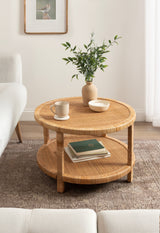 Round two-tier rattan coffee table with light brown woven texture, decorated with a beige vase holding leafy branches, a white cup, and a small bowl.