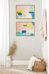 Two abstract paintings with soft pastel backgrounds featuring bold blue, yellow, and red brushstrokes, framed in simple wooden frames and hung vertically on a light gray wall.