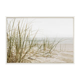 Photograph of soft beige sand dunes with sparse green beach grass under a pale overcast sky, framed by a simple light wood frame.
