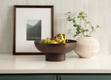 Dark bronze metal bowl with vertical ribbed base holding artichokes, placed on a cream countertop next to a ribbed white vase with green leafy stems and a framed mountain photo.