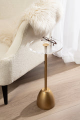 Small round side table with clear glass top and brass-colored pedestal base, next to a white textured armchair with a fluffy white throw, on light wood flooring.