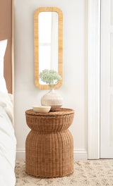 Tall rectangular mirror with rounded corners and a textured wooden frame, mounted on a white wall above a woven wicker stool topped with a striped ceramic vase and bowl.