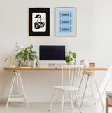Minimal wooden desk with white trestle legs, two potted plants, a white desk lamp, and an iMac below framed art of cherries with 8-balls on black and blue Cheers tickets.