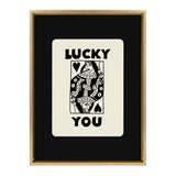 Black and white graphic resembling a playing card with a stylized queen figure, titled 'Lucky You' in bold uppercase letters, framed in natural wood.
