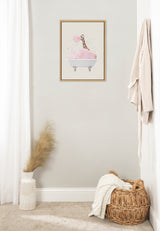 Framed print of a giraffe blowing a pink bubble in a clawfoot bathtub filled with pink bubbles, hanging on a light gray wall above a woven basket and a white vase with pampas grass.